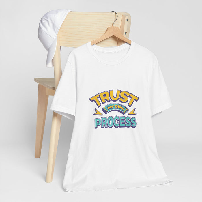 A white short-sleeve t-shirt on a wooden hanger, styled in front of a light wood chair with a white cap draped over the back. The shirt features a colorful retro-style graphic on the chest reading 'TRUST MY OWN PROCESS" | AH Urban Wear PK