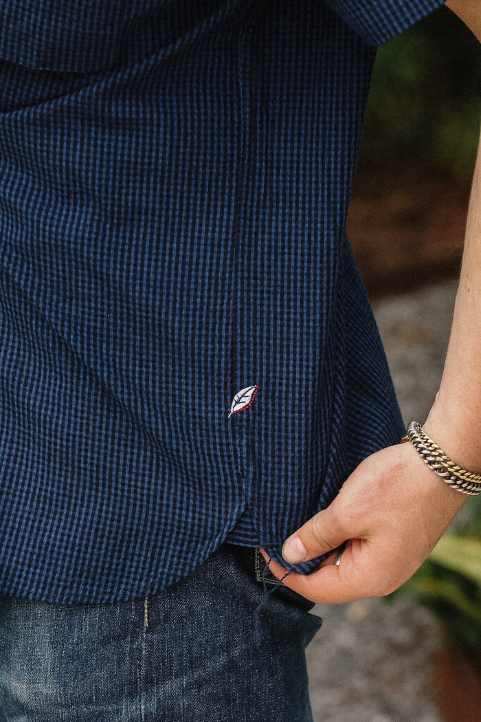 Close-up of a person wearing a navy blue gingham check short-sleeve button-down shirt, pulling the hem slightly with a hand. A small embroidered logo is visible on the lower front, and the person is also wearing blue jeans and a chain bracelet.