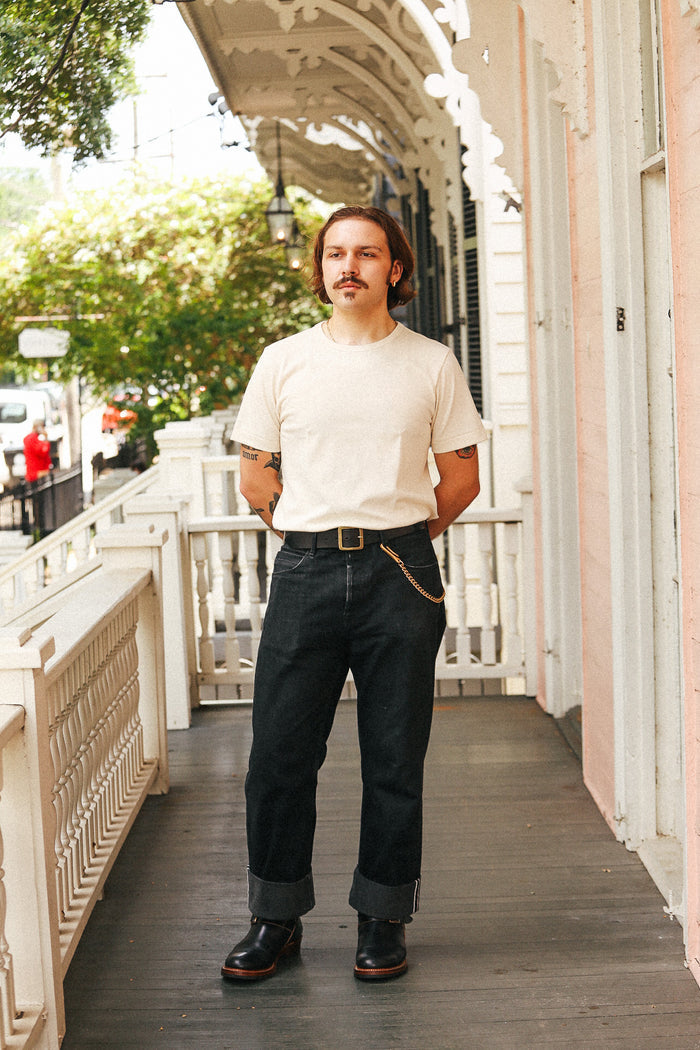 A man stands on a porch wearing a cream crewneck t-shirt, dark blue jeans with rolled cuffs, a brown leather belt with a gold buckle, and black leather shoes.