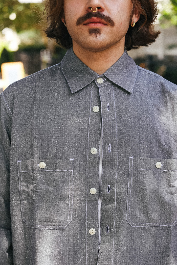 A close-up of a man wearing a gray chambray button-down shirt with two chest pockets and white buttons, set against a blurred outdoor background.