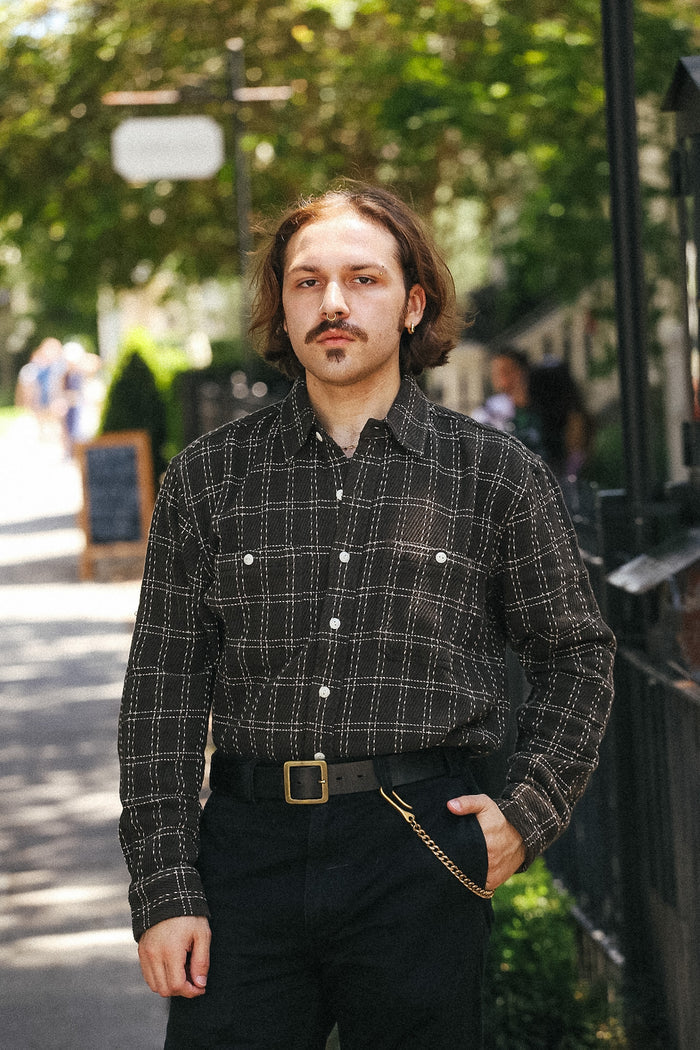 A man wearing a black and brown plaid button-down shirt with a gold chain detail at the waist, standing outdoors on a sunny day.
