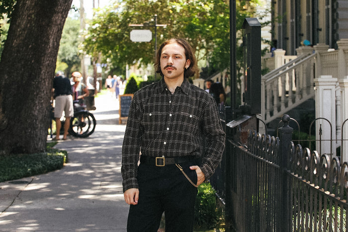 A man stands on a sunlit sidewalk wearing a dark plaid button-up shirt and black pants, with trees, a house, and a fence in the background.