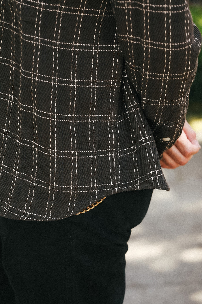 Close-up of a person wearing a dark brown plaid overshirt with white stitched windowpane lines and black pants, standing outdoors.