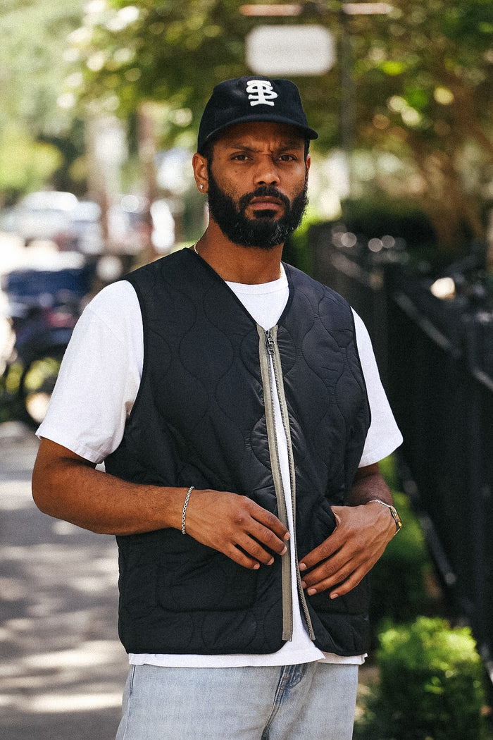 A man wearing a black quilted zip-up vest over a white t-shirt, light blue jeans, and a black baseball cap with a white logo, standing outdoors on a blurred street background.