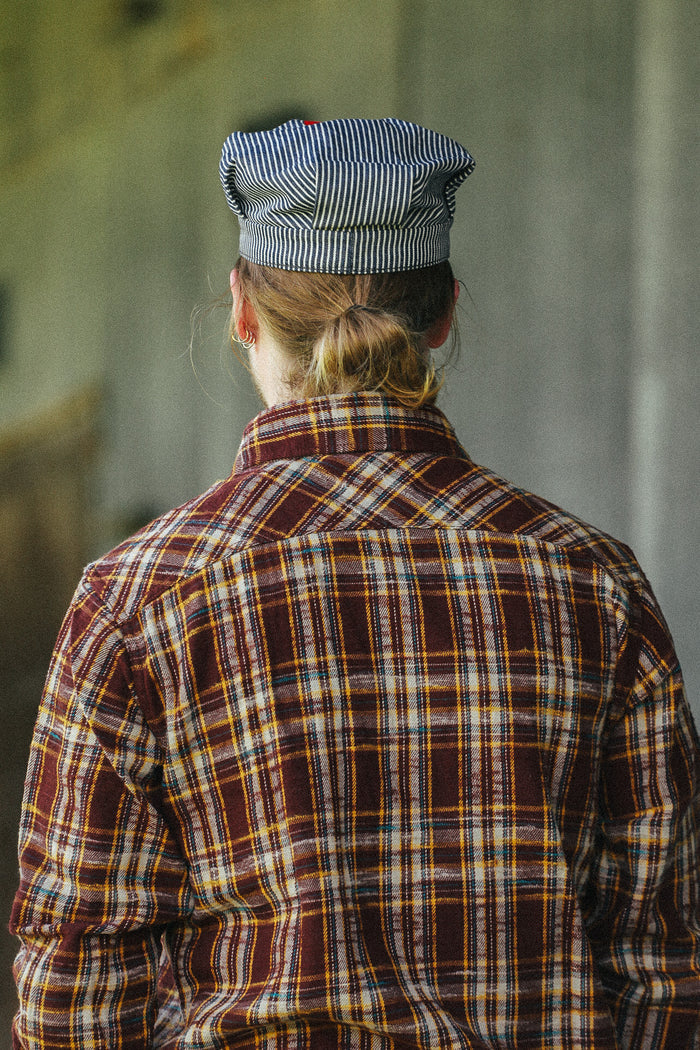 A person viewed from behind wearing a brown and red plaid flannel shirt with yellow and white stripes, and a blue-and-white striped cap, standing against a blurred gray background.