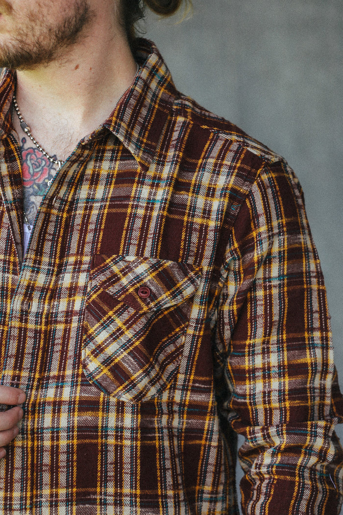 A close-up of a man wearing a brown, yellow, and black plaid button-down shirt, showing the chest pocket and collar area.