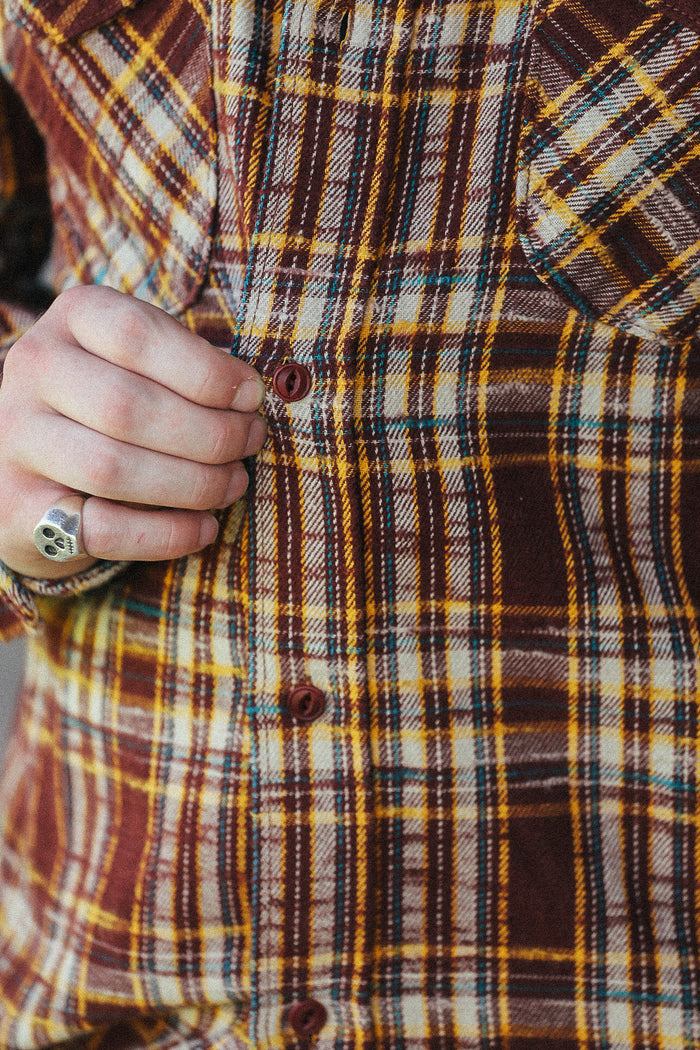 Close-up of a person wearing a brown, yellow, and cream plaid button-up shirt, with a hand pulling the front button.
