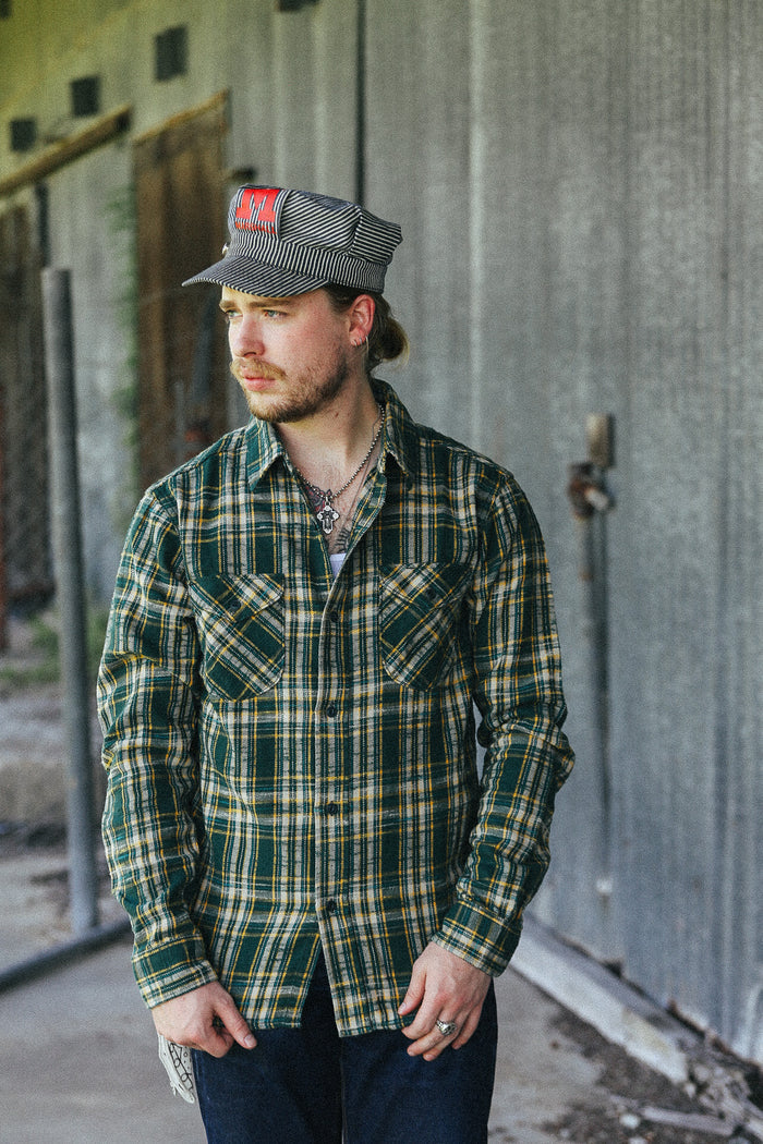 A man wearing a green and yellow plaid button-down shirt, dark jeans, and a gray striped cap with a red patch, standing in front of a corrugated metal wall.