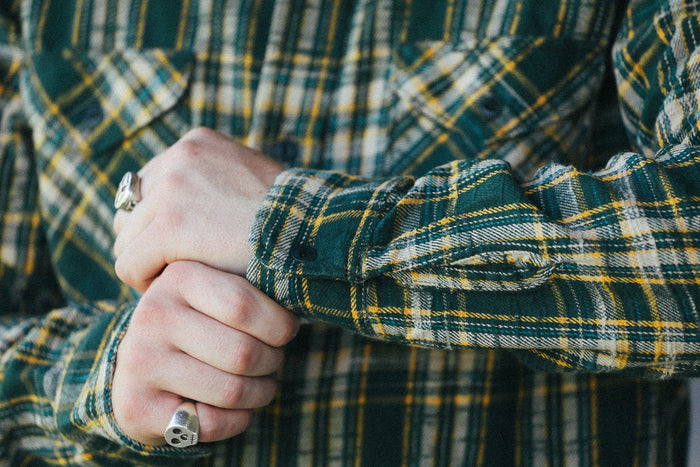 A close-up of a person's hands clasped in front of a green and yellow plaid flannel shirt. The person is wearing silver rings on both fingers.