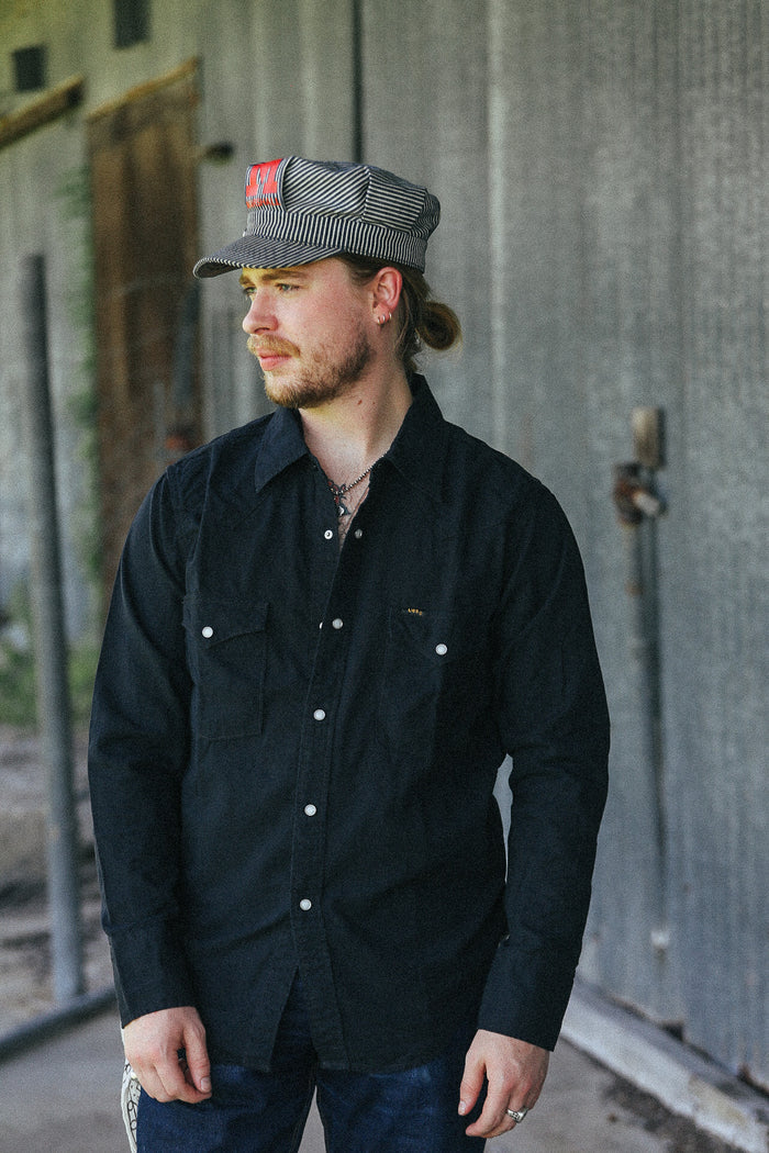 A man wearing a black long-sleeve button-up shirt with silver snap buttons and chest pockets, paired with a gray and black striped cap, standing in front of a gray corrugated metal wall.