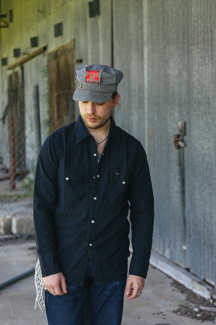 A man wearing a dark navy button-up shirt, blue jeans, and a gray striped cap with a red square patch, standing in front of a weathered concrete wall.