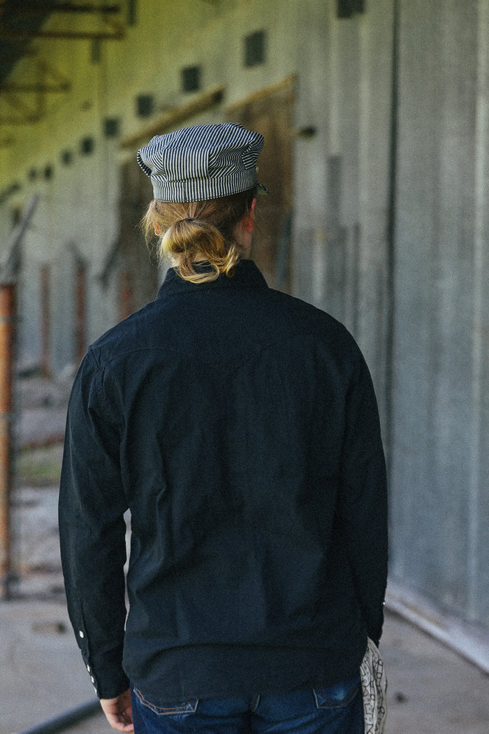 A person viewed from behind wearing a dark navy long-sleeve shirt and a gray-and-white striped cap, standing in a corridor with a concrete wall.