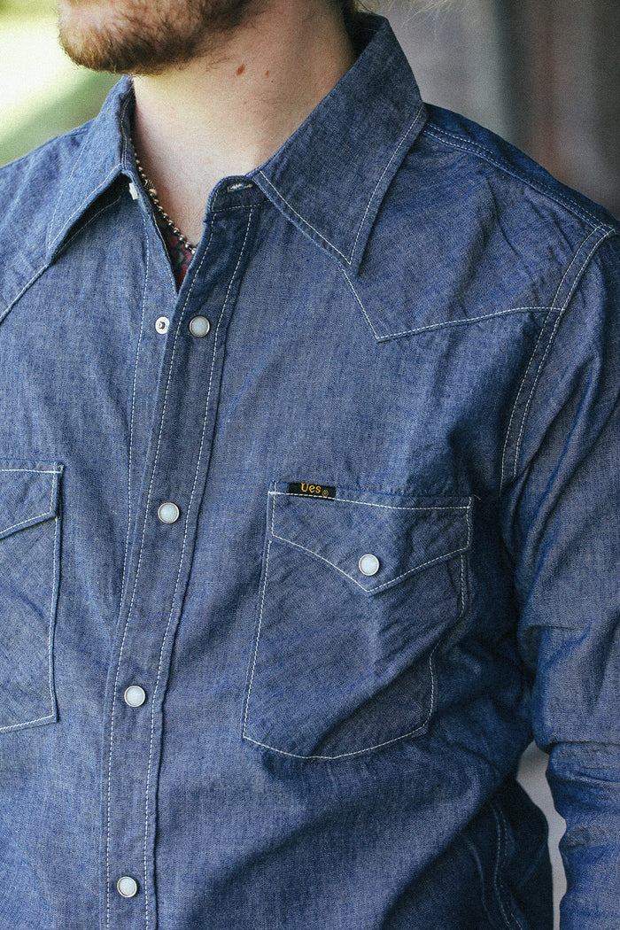 A close-up of a man wearing a blue denim western-style button-up shirt with a chest pocket and a small yellow brand tab.