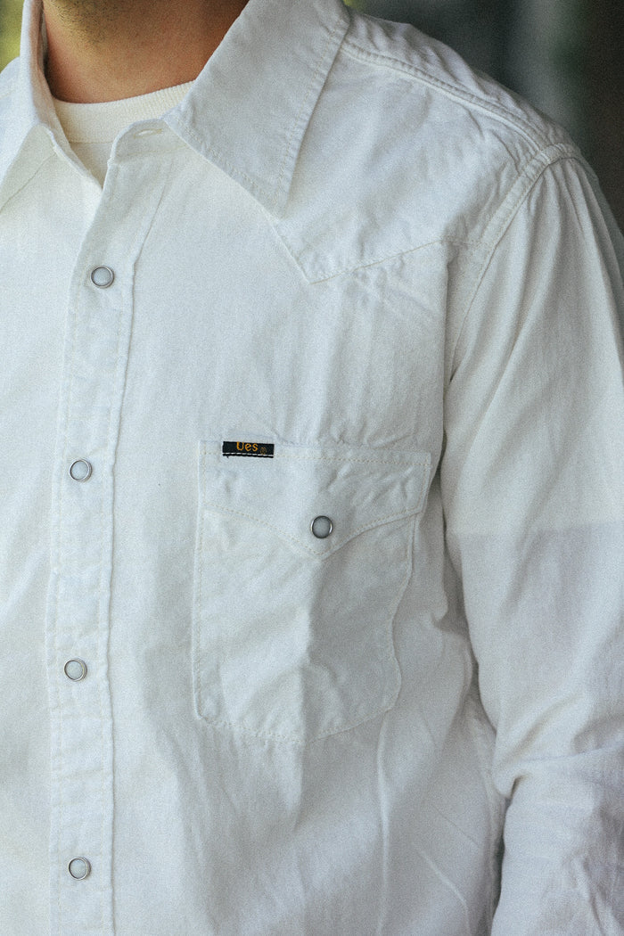 A close-up of a person wearing a white long-sleeve button-up shirt with a chest pocket and a small black brand label.