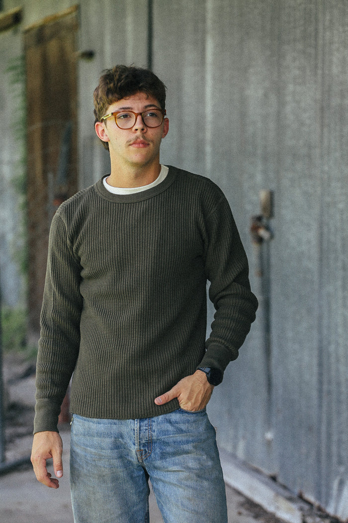 A young man wearing a dark olive green ribbed knit sweater over a white undershirt and blue jeans, standing in front of a weathered gray metal wall.