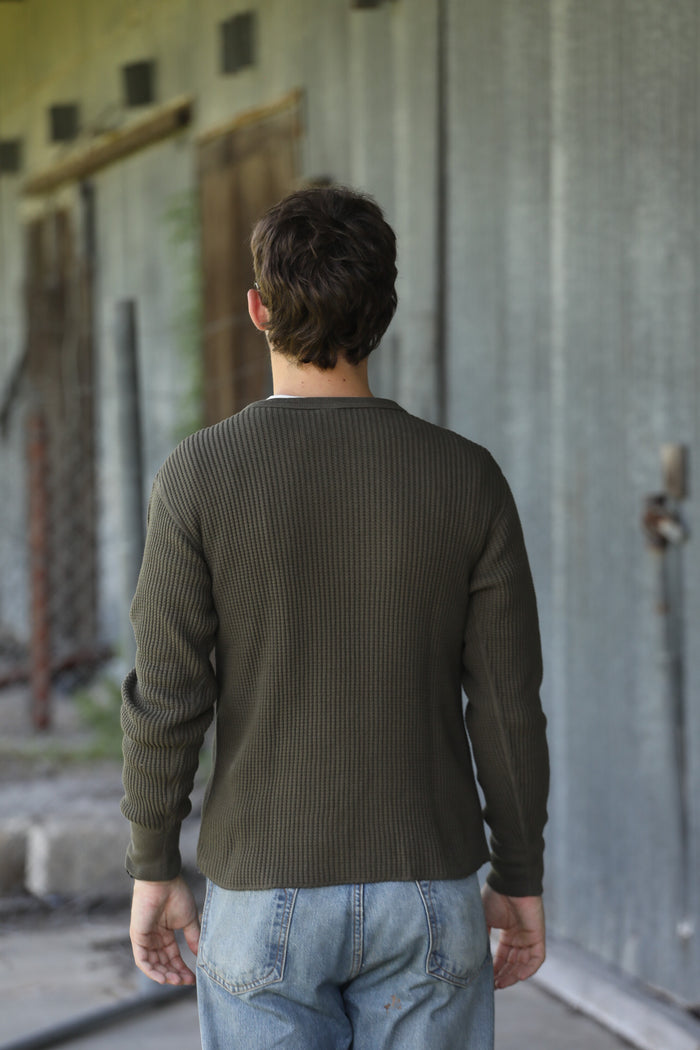 A man viewed from behind wearing an olive green ribbed long-sleeve shirt and light blue jeans, standing in front of a weathered gray metal wall.