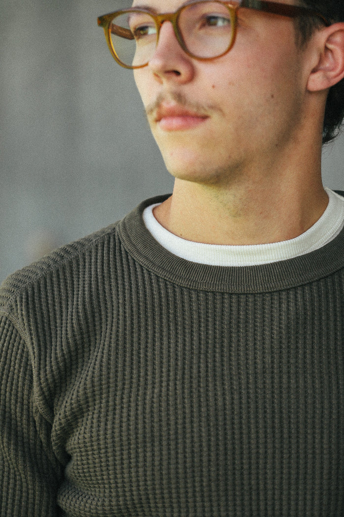 A close-up shot of a man wearing a dark olive green ribbed knit crewneck sweater over a white t-shirt, with brown-rimmed eyeglasses, set against a soft gray background.