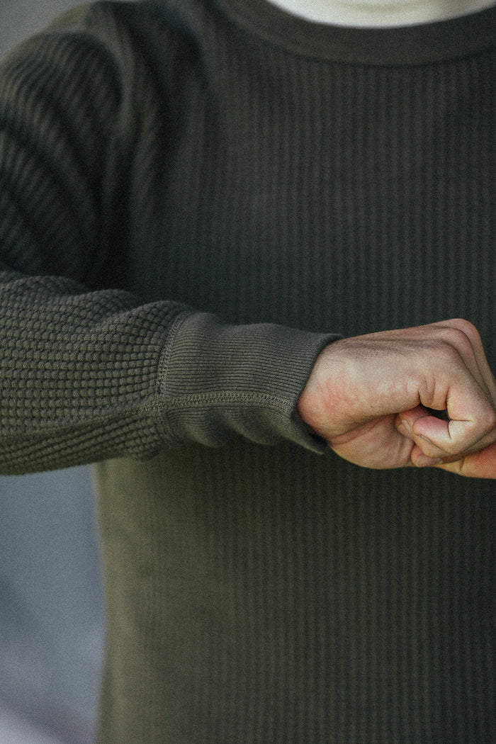 Close-up of a person wearing a dark olive green ribbed knit sweater, showing the sleeve cuff and hand.