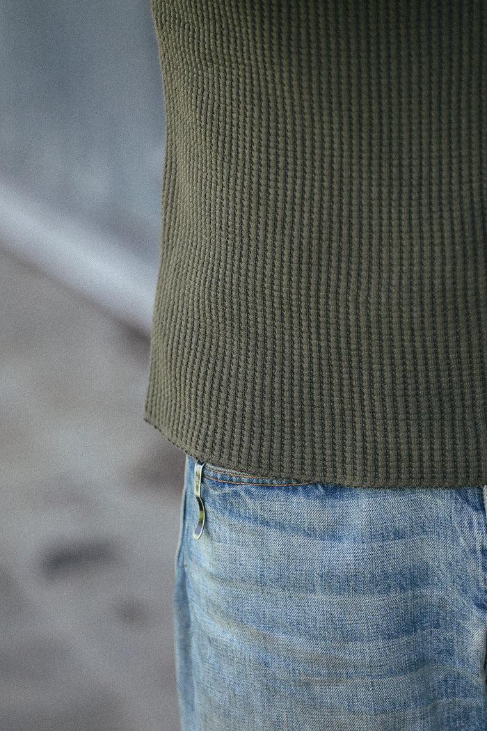 Close-up of a person wearing an olive green ribbed knit top and light blue jeans, cropped from the chest down against a blurred gray background.
