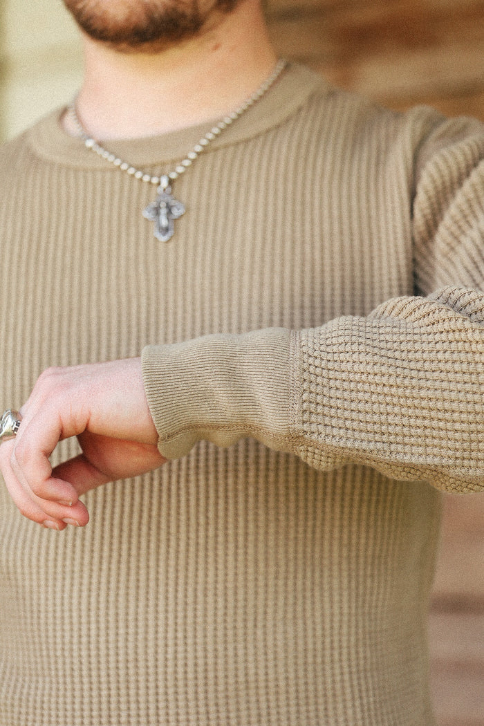 A close-up of a man wearing a beige ribbed knit sweater, a silver beaded necklace with a cross pendant, and a silver ring.
