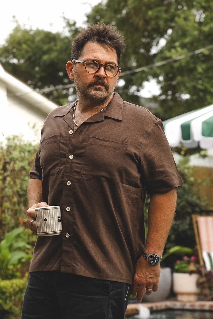 A man wearing a brown short-sleeve button-up shirt and glasses, holding a white mug in an outdoor backyard setting.