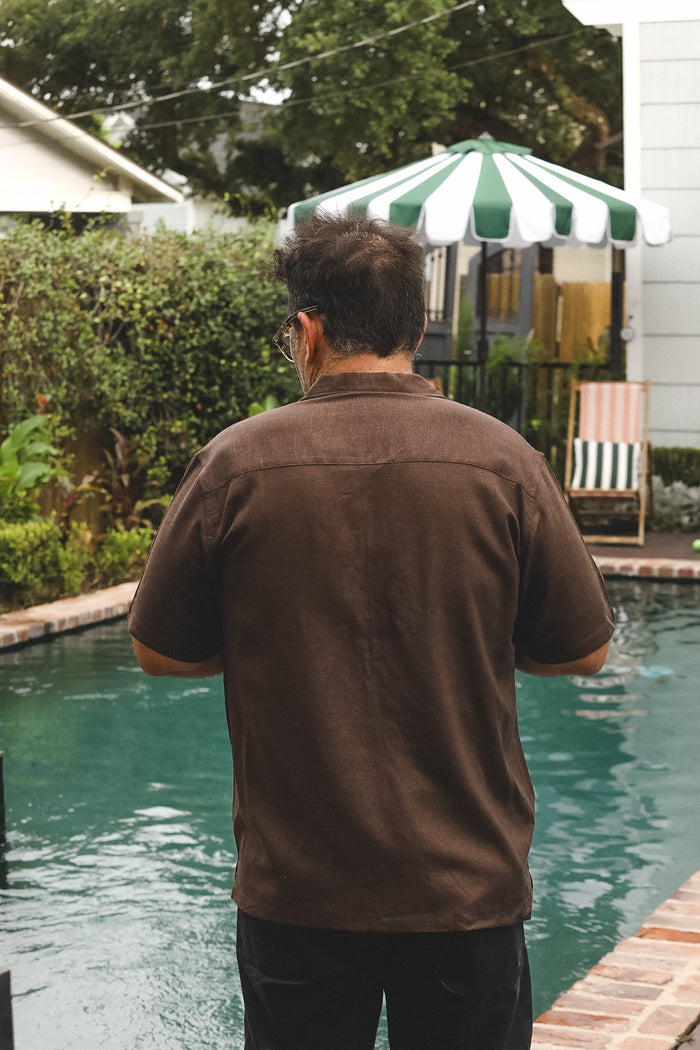 A man viewed from behind wearing a dark brown short-sleeve button-up shirt and black pants, standing by a swimming pool in a backyard with a green and white striped patio umbrella and garden foliage in the background.