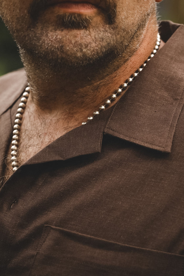 A close-up shot of a man wearing a silver beaded necklace over a brown collared shirt.
