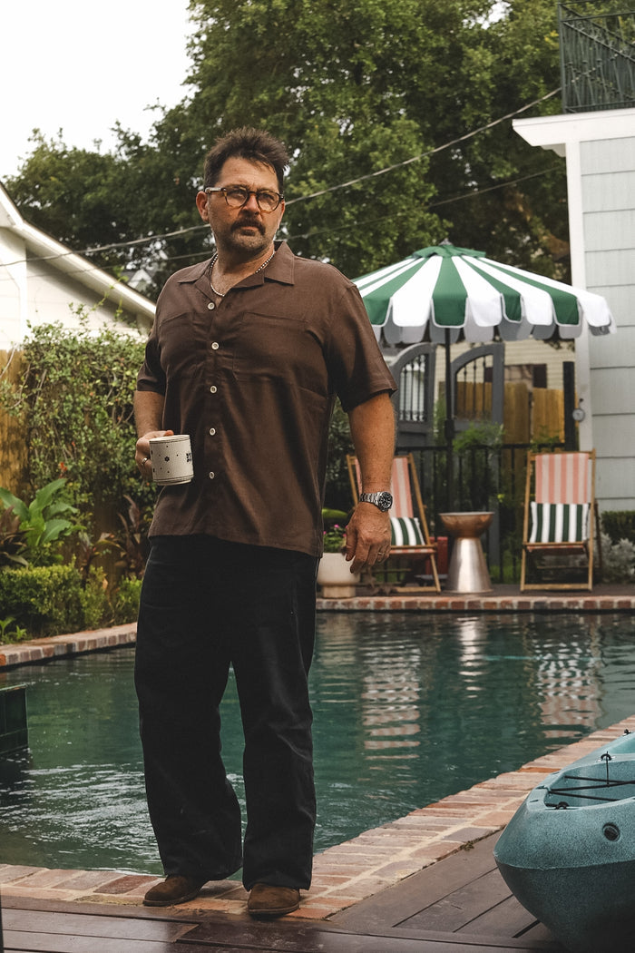 A man wearing a brown short-sleeve button-up shirt and black pants stands by a swimming pool holding a white mug, with a green and white striped patio umbrella and lounge chairs in the background.