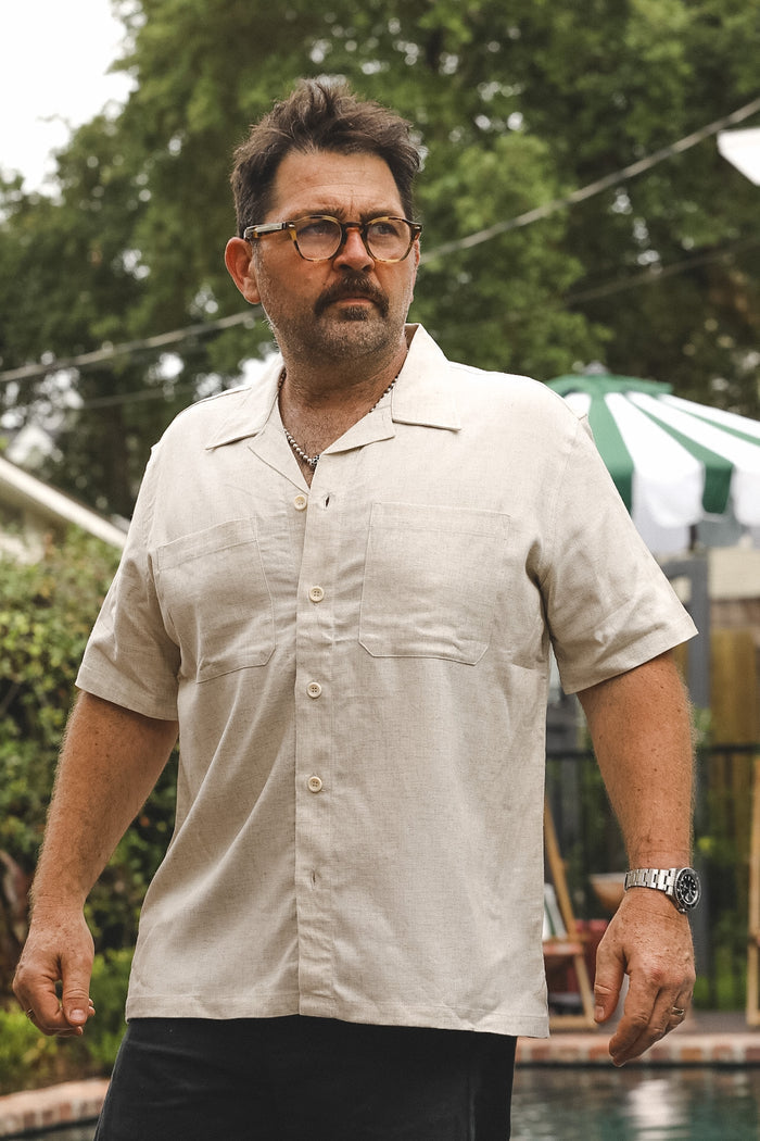 A man wearing a light beige short-sleeve button-up shirt and black pants, standing outdoors near a pool with trees and patio furniture in the background.