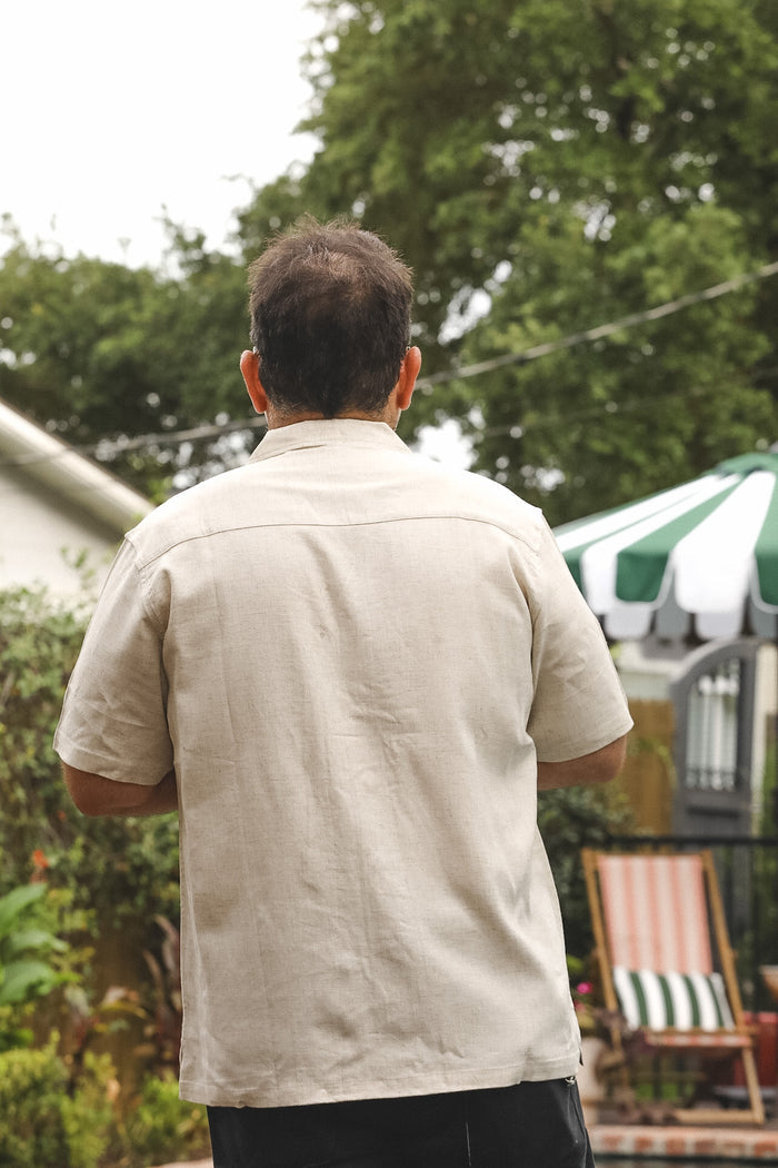 A man viewed from behind wearing a light beige short-sleeve linen shirt and dark pants, standing in an outdoor backyard setting with trees, a patio umbrella, and a chair in the background.