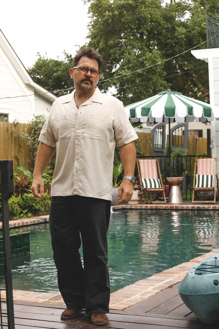 A man wearing a beige short-sleeve button-up shirt and dark pants stands on a wooden deck next to a swimming pool with patio chairs and a green-and-white striped umbrella in the background.