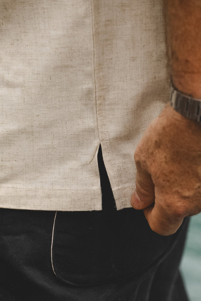 Close-up of a person wearing a light beige linen shirt with a front slit hem, paired with black pants and a dark wristwatch.