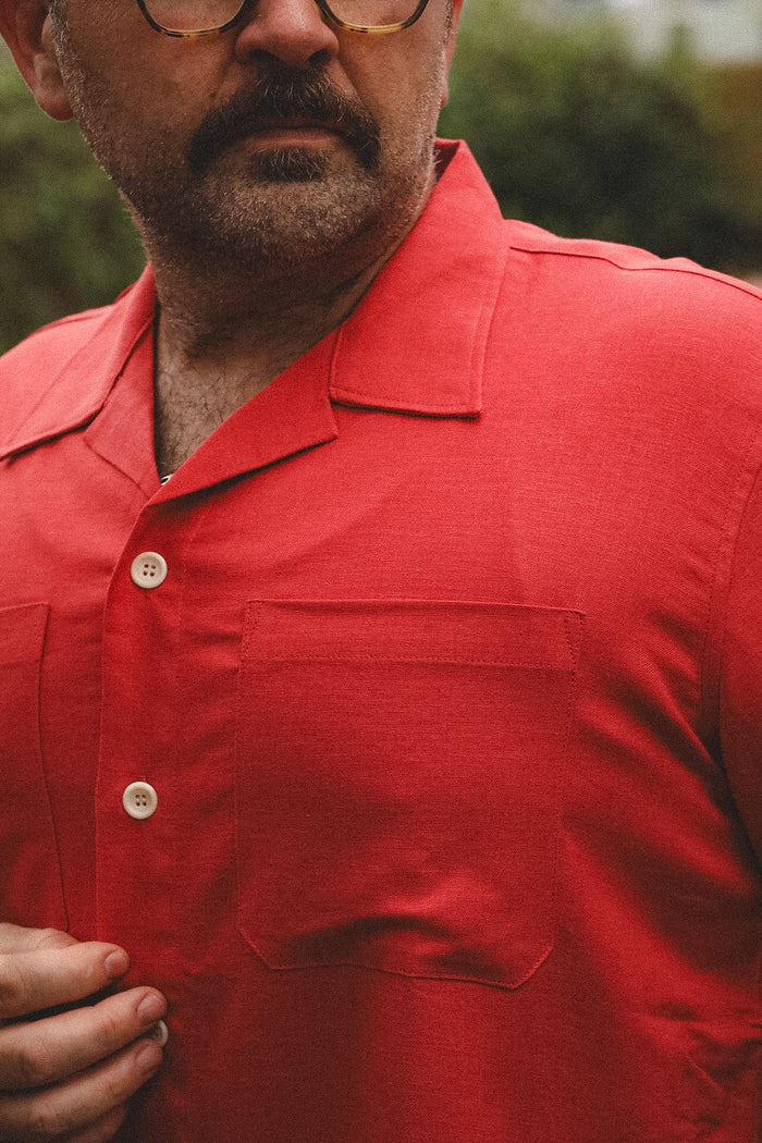 A close-up of a man wearing a bright red short-sleeve camp collar button-up shirt with a chest pocket, set against a blurred outdoor background.