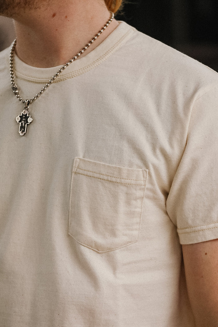 Person wearing a beige t-shirt with a pocket and a necklace with a pendant.