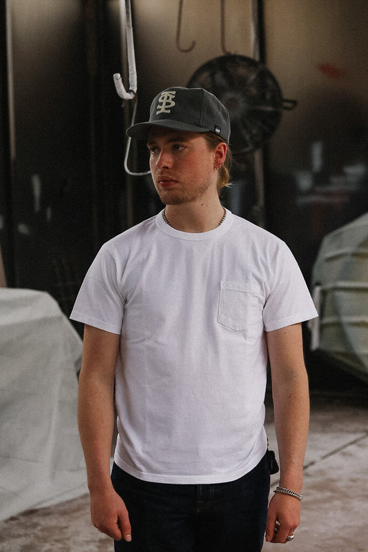 Person wearing a white t-shirt and dark cap, standing in front of a reflective surface.