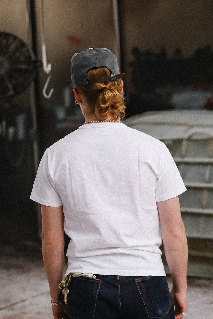 Person wearing a white t-shirt and dark cap, standing in front of a reflective surface.