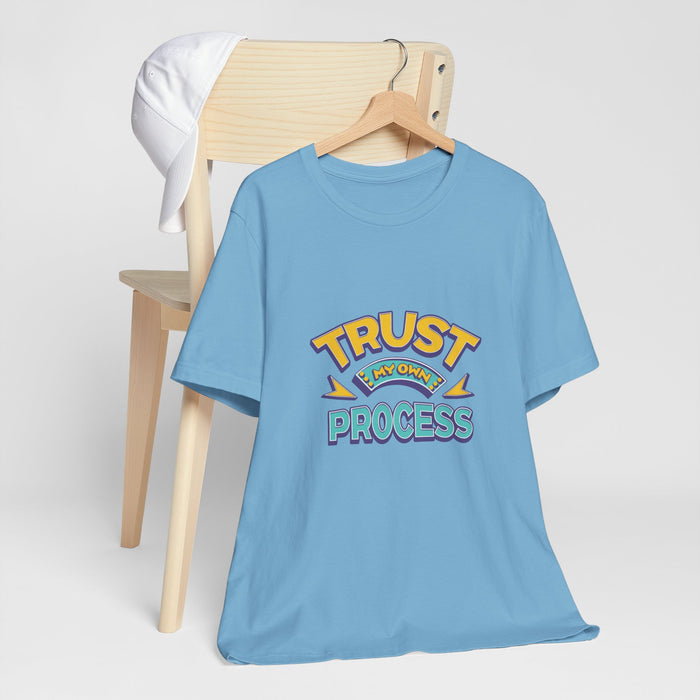 A Women light blue short-sleeve t-shirt with a graphic reading 'TRUST MY OWN PROCESS' in yellow, ,and teal, hanging on a wooden hanger from a light wood chair with a white shirt draped over it, against a light gray background | AH Urban Wear PK