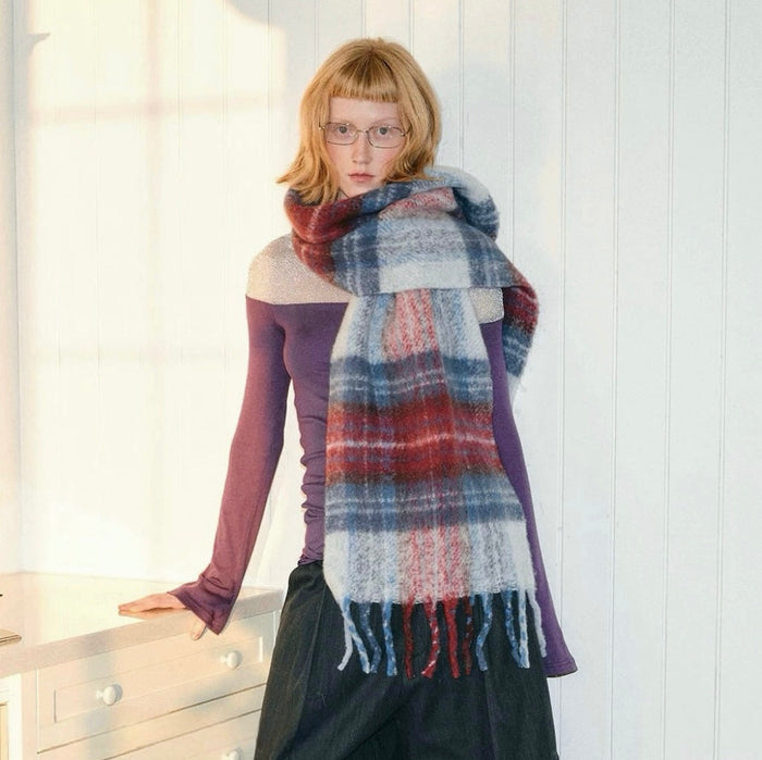 Model wearing Midnight Blaze Plaid Scarf, a long wool blend scarf with red, blue, and white plaid pattern and fringe.
