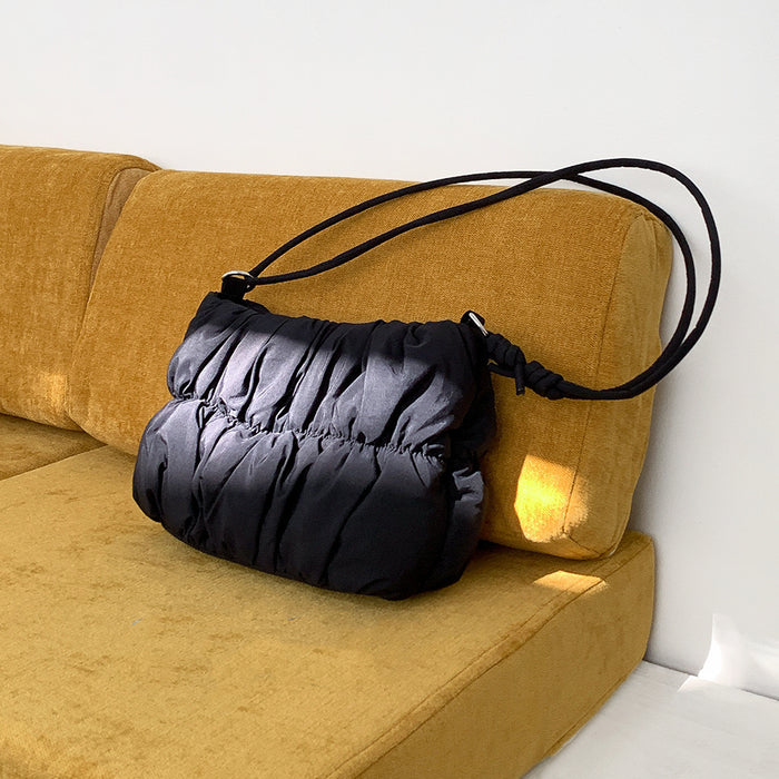 Front view of Black pleated  cloud bag with an adjustable strap, resting on a yellow couch Pleated Cloud.