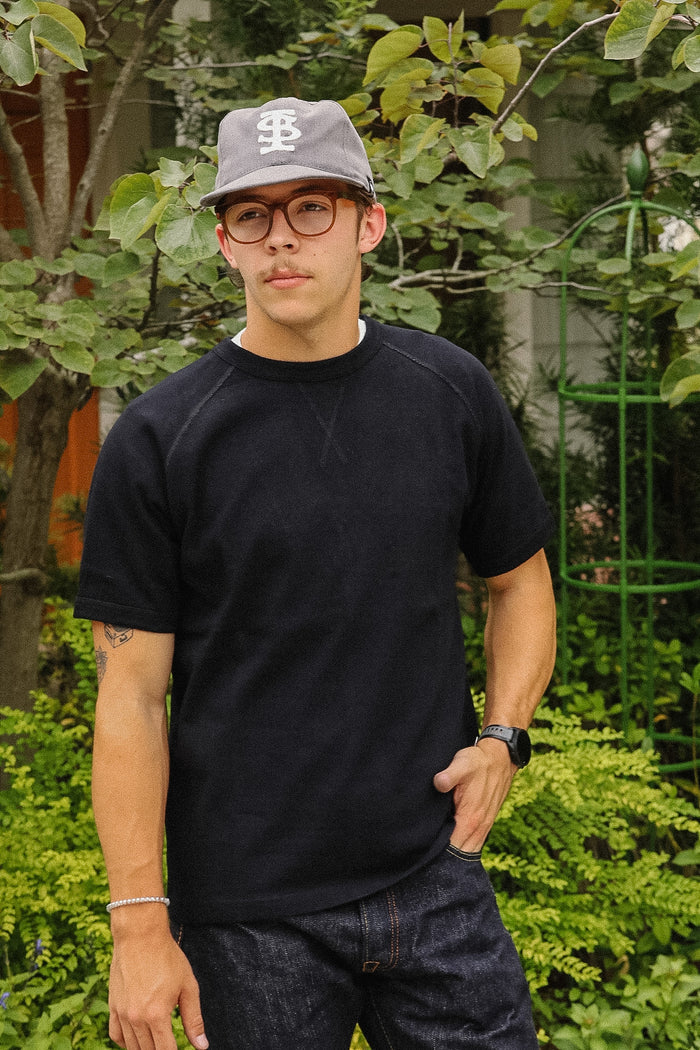 A man wearing a black short-sleeve crewneck sweatshirt, dark denim jeans, a gray baseball cap with a white 'BS' logo, and glasses, standing outdoors in front of green foliage.
