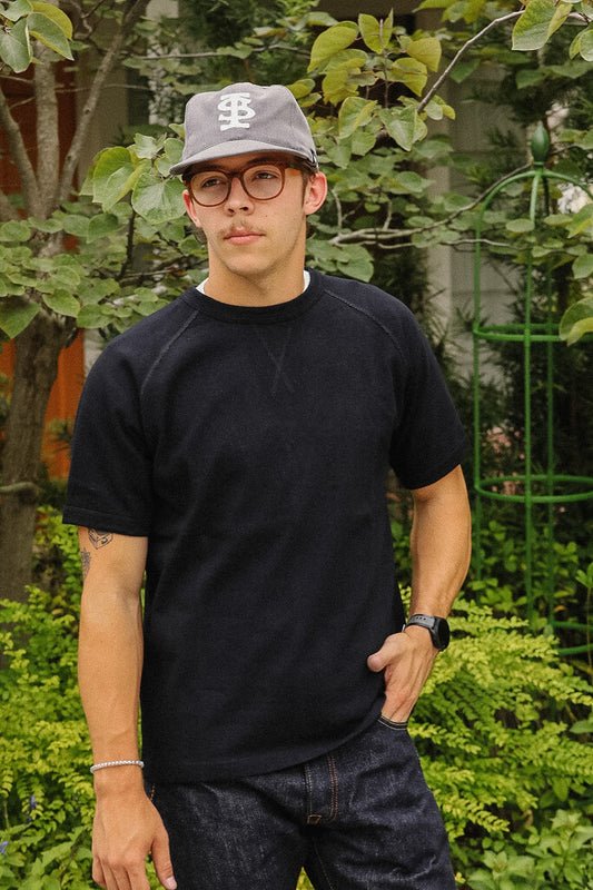 A man wearing a black short-sleeve crewneck sweatshirt, dark denim jeans, a gray baseball cap with a white 'BS' logo, and glasses, standing outdoors in front of green foliage.
