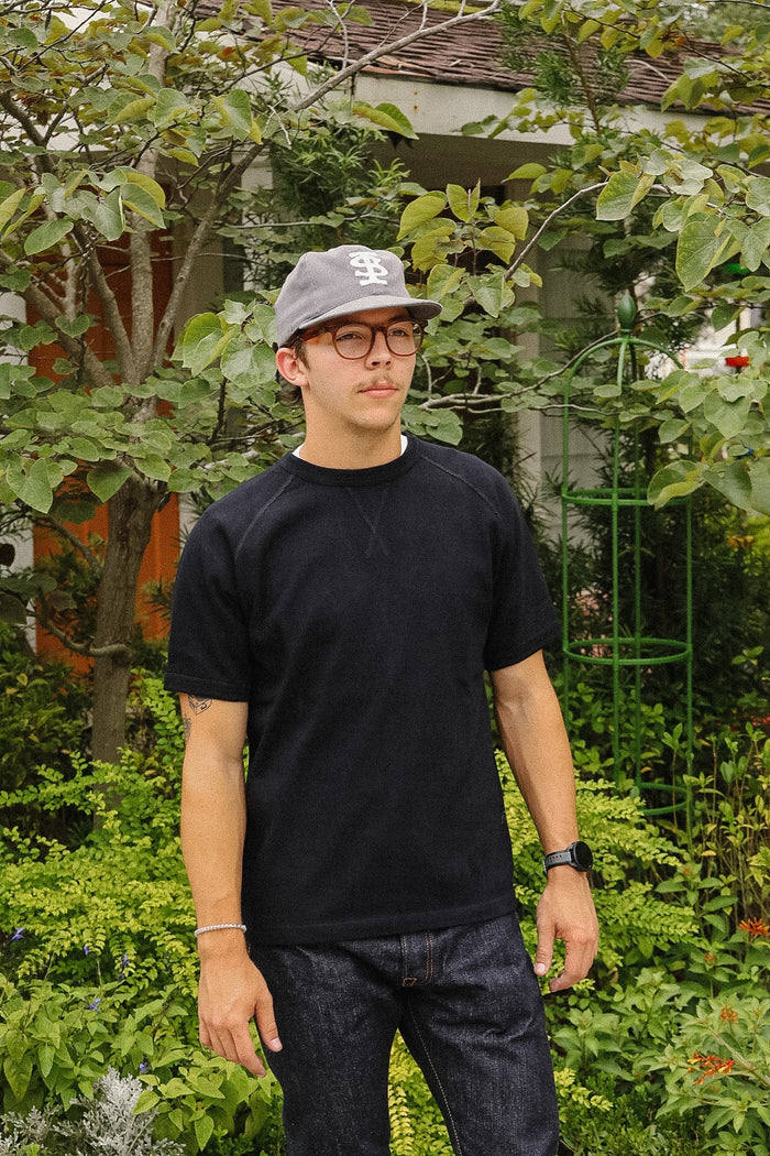 A man wearing a black crewneck t-shirt, dark jeans, a gray baseball cap, and glasses, standing in a lush green garden setting.