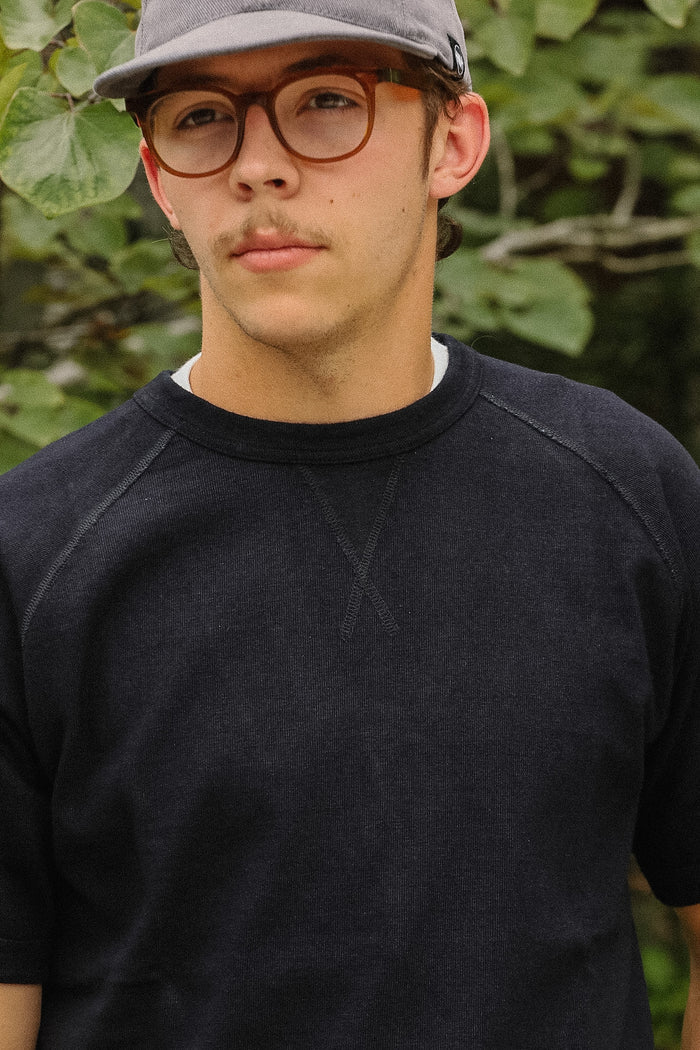 Close-up of a person wearing a dark charcoal gray ribbed crewneck t-shirt, with a blurred green garden background.