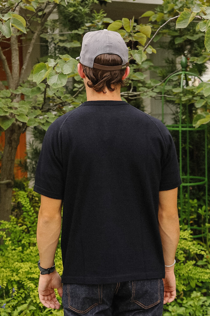 A man viewed from behind wearing a dark navy blue short-sleeve t-shirt, a gray baseball cap, and dark jeans, standing outdoors in front of green foliage.