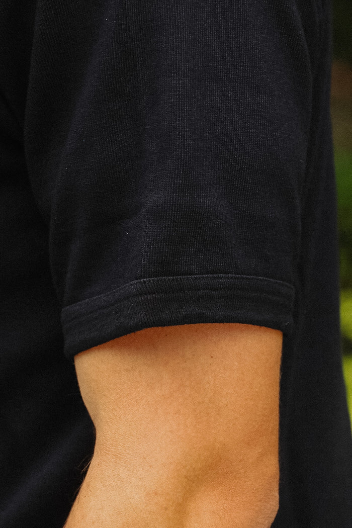 Close-up of a person's forearm wearing a black short-sleeve ribbed knit top, showing the sleeve cuff and skin against a blurred green background.