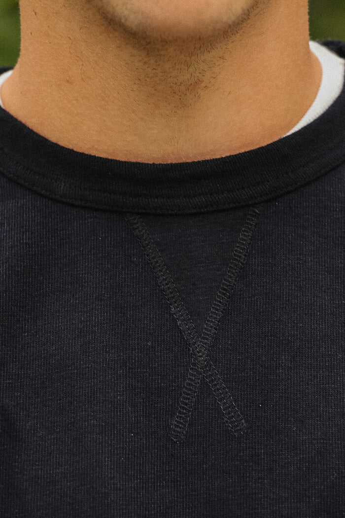 Close-up of a person wearing a dark charcoal crewneck sweatshirt, showing the ribbed collar and the crossed-knit chest detail.