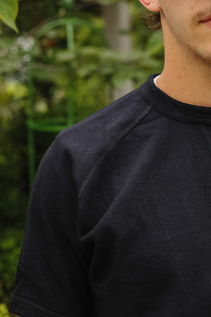 Close-up of a person wearing a dark charcoal gray ribbed crewneck t-shirt, with a blurred green garden background.
