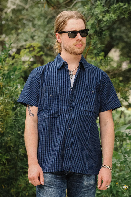 A man wearing a navy blue short-sleeve button-up shirt with a subtle vertical stripe pattern, styled with blue jeans, sunglasses, and a necklace, standing outdoors in front of a green foliage background.
