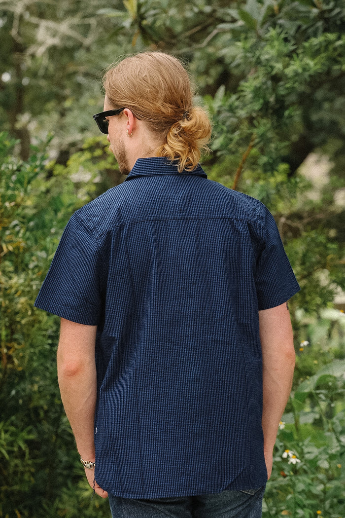 A man with blonde hair tied back and wearing sunglasses is shown from behind, wearing a dark navy blue short-sleeve button-down shirt with a subtle grid pattern, standing outdoors against a background of green foliage.