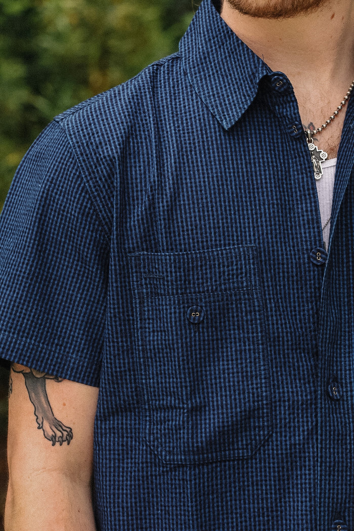Close-up of a person wearing a short-sleeve button-up shirt with a fine blue and white micro-check pattern, featuring a chest pocket and visible tattoos on the arm.